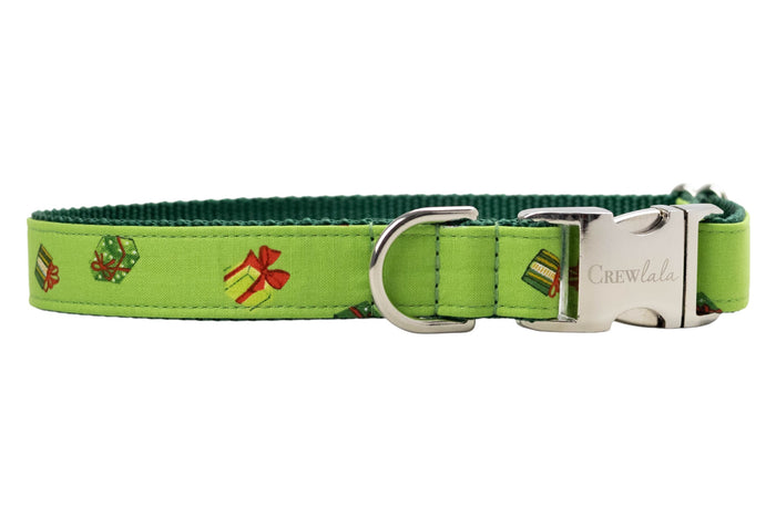 Festive dog collar made from bright green fabric printed with small colorful gift boxes in festive colors, each tied with decorative bows. The collar features dark green woven backing for durability and metal hardware, including a branded “Crewlala” buckle.