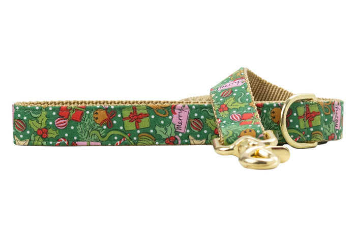 Festive green dog leash decorated with small holiday icons including presents, holly, candy canes, bells, and ornaments, over a dotted background. Nylon webbing underside and hardware add a warm seasonal touch.