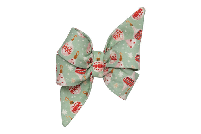 An oversized dog belle bow made from light green fabric patterned with vintage Christmas ornaments, hooks, and scattered snowflakes. The bow features large, structured loops and long, pointed tails, giving it a dramatic, festive appearance—perfect for attaching during the holiday season