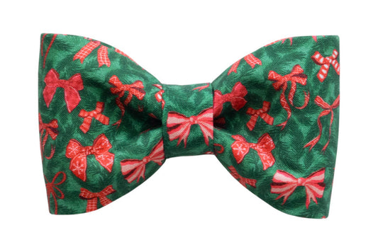 A festive dog bow tie made from green fabric with an all-over print of bows, candy canes, and ribbon designs. The bow tie has a symmetrical, classic shape with a gathered center and is designed to be a cheerful holiday look.