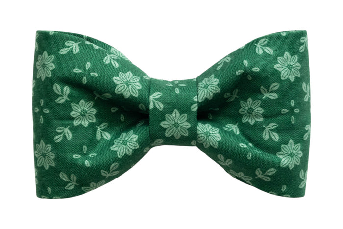 A classic dog bow tie made from green fabric with a light green floral pattern featuring symmetrical flowers and scattered leaf shapes. The bow tie has a crisp, structured shape with a cinched center and is designed to attach to a collar for a simple, elegant look.