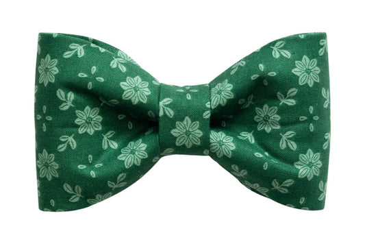 A classic dog bow tie made from green fabric with a light green floral pattern featuring symmetrical flowers and scattered leaf shapes. The bow tie has a crisp, structured shape with a cinched center and is designed to attach to a collar for a simple, elegant look.
