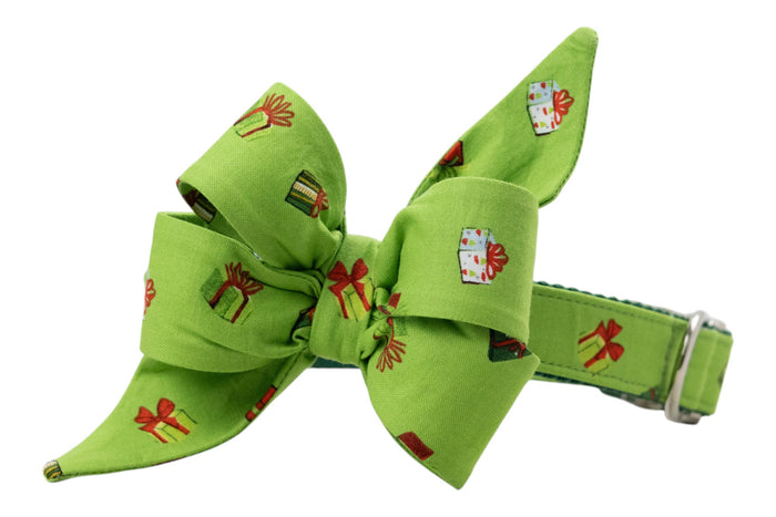 Festive dog collar in light green with a large, structured belle bow attached, both covered in a whimsical print of colorful wrapped presents. The feminine belle bow features exaggerated loops and pointed ends for a playful holiday look, and the collar includes an adjustment buckle.