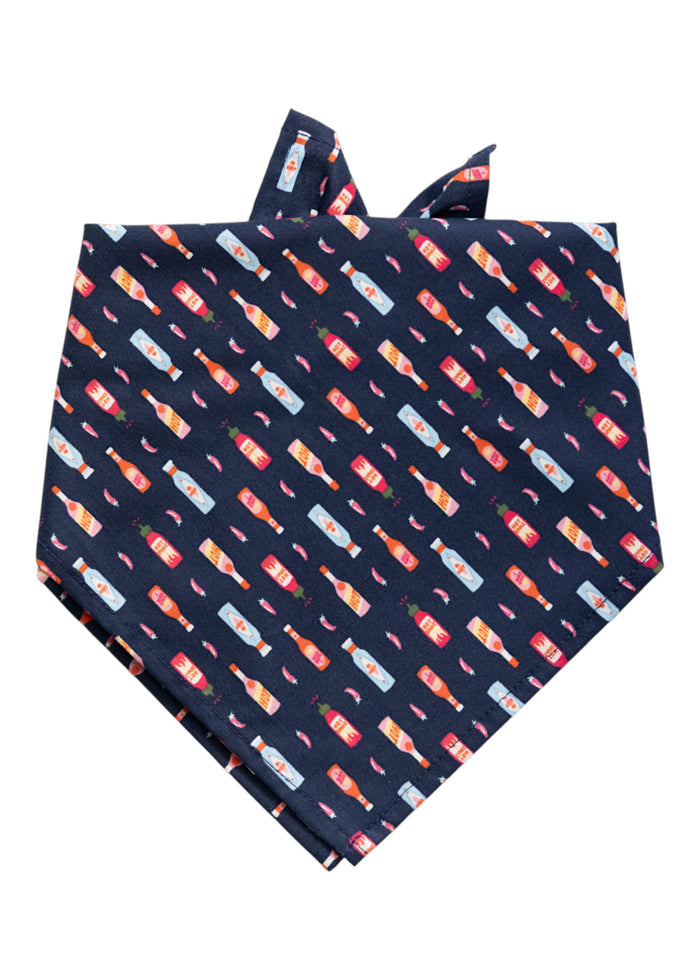 Dark navy blue dog bandana decorated with a repeating diagonal pattern of illustrated hot sauce bottles in an array of colors, each with small label details, interspersed with tiny chili peppers.