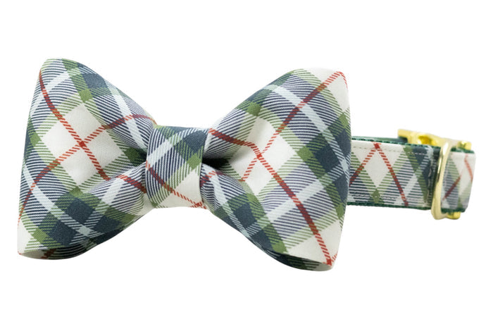 Dog collar with attached, classic bow tie in light fabric featuring a tartan plaid pattern, accented with fine diagonal stripes. The bow tie has a symmetrical, structured shape and is fixed to the matching plaid collar, which includes a polished D-ring and adjustable buckle.