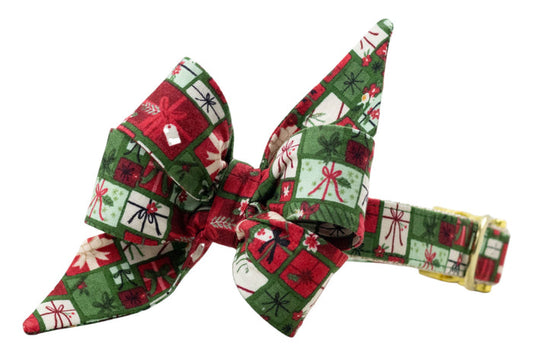 Oversized dog belle bow collar with festive gift box pattern, featuring bows, ribbons, holly, and tags. The belle bow has large pointed tails and is attached to a matching collar with polished hardware.