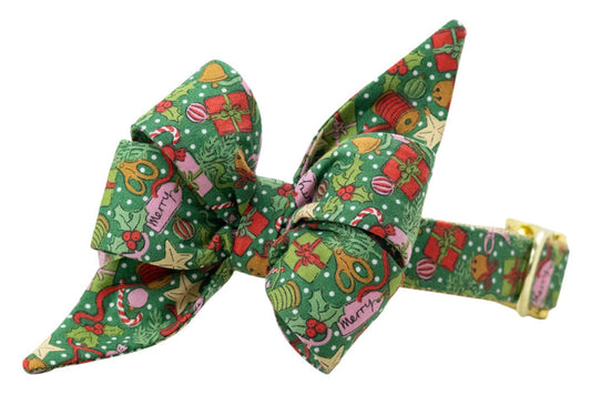 Festive green dog collar with a decorative,  matching belle bow, covered in a colorful holiday print of gifts, holly, bells, candy canes, and ornaments. Finished with shiny hardware for a polished, seasonal look.