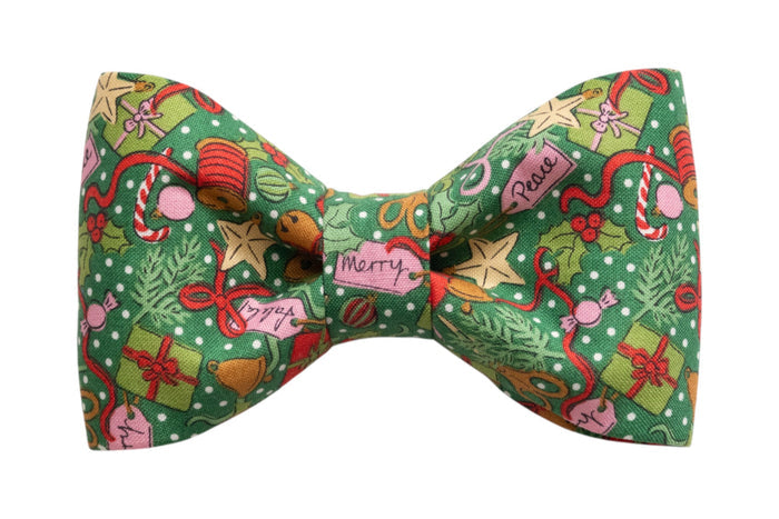 Green fabric bow tie patterned with colorful Christmas icons including gifts, candy canes, stars, holly, and holiday tags reading “Merry” and “Peace,” perfect for your dog's festive winter look.