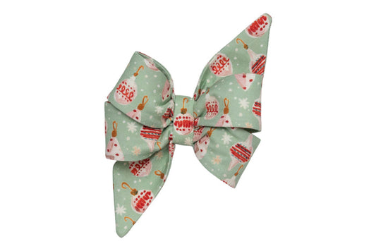 An oversized dog belle bow made from light green fabric patterned with vintage Christmas ornaments, hooks, and scattered snowflakes. The bow features large, structured loops and long, pointed tails, giving it a dramatic, festive appearance—perfect for attaching during the holiday season