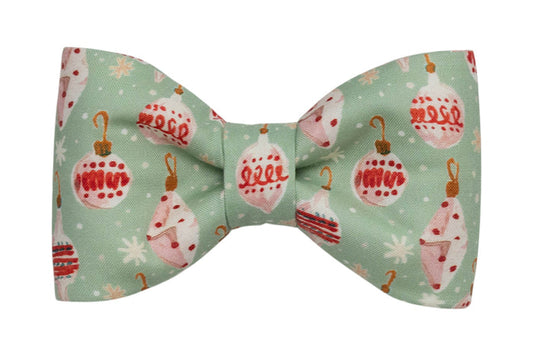 Holiday-themed dog bow tie in soft green fabric featuring illustrated Christmas baubles in festive colors, surrounded by snowflake and polka dot accents. The bow is centered and neatly cinched for a tidy, classic appearance, ideal for seasonal wear.